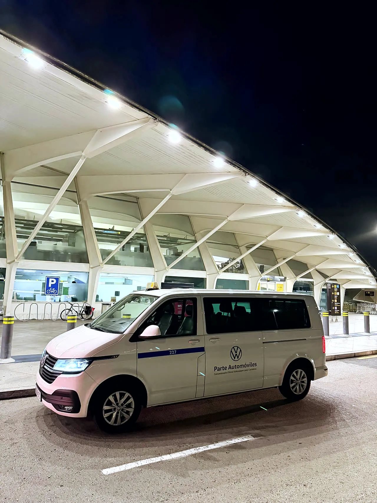 Furgoneta blanca de 8 plazas junto a la terminal de un aeropuerto por la noche